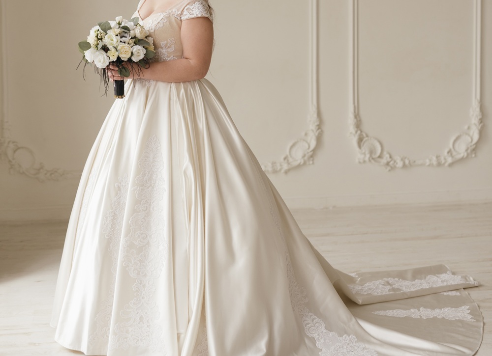 A person in a white wedding dress holds a bouquet of flowers, standing in an elegant, light-colored room with decorative wall molding.