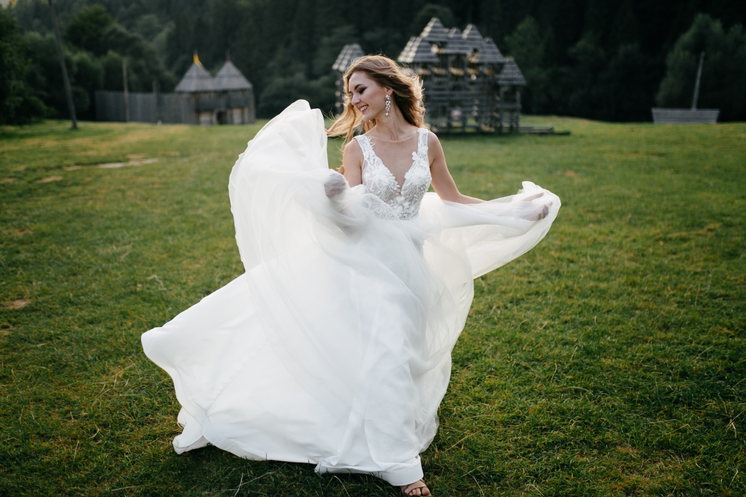 A woman in a white wedding dress twirls on grass in an open field, with wooden structures and trees in the background.